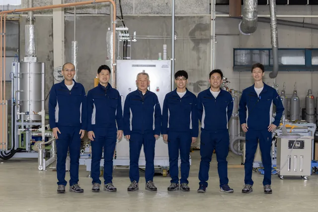 岐阜工場のメンバー。作業着姿の8人が量産機の前に並んでいる。
